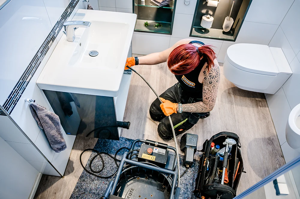 Eine Mitarbeiterin von Schoenen Abwassertechnik mit roten Haaren und Tätowierungen, die orangefarbene Handschuhe trägt, führt mit einer Inspektionsausrüstung eine Rohrreinigung unter einem Waschbecken im Badezimmer eines Hauses in Köln durch. Werkzeuge und Maschinen liegen in einem modernen Badezimmer auf dem Boden.