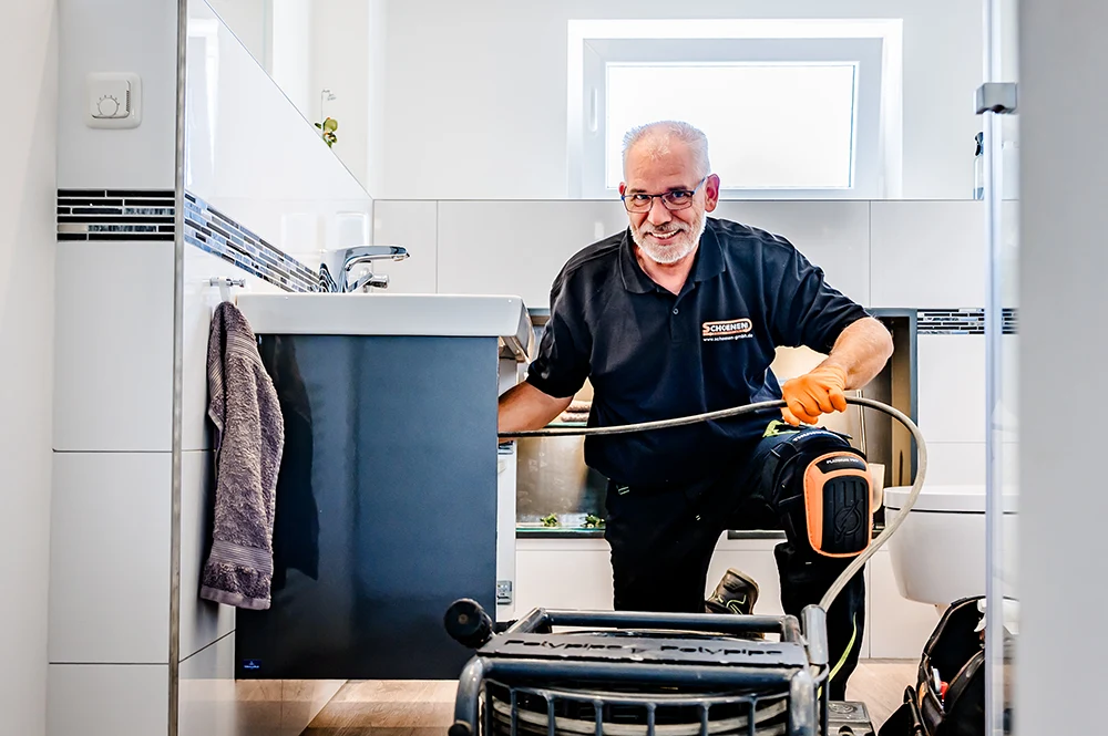 Ein Mitarbeiter mit Handschuhen und Knieschonern arbeitet mit professionellem Werkzeug unter einem Waschbecken im Badezimmer, da der Abfluss verstopft ist. Er lächelt in die Kamera. Das Badezimmer ist modern mit weißen Fliesen und einem Handtuch, das in der Nähe hängt.