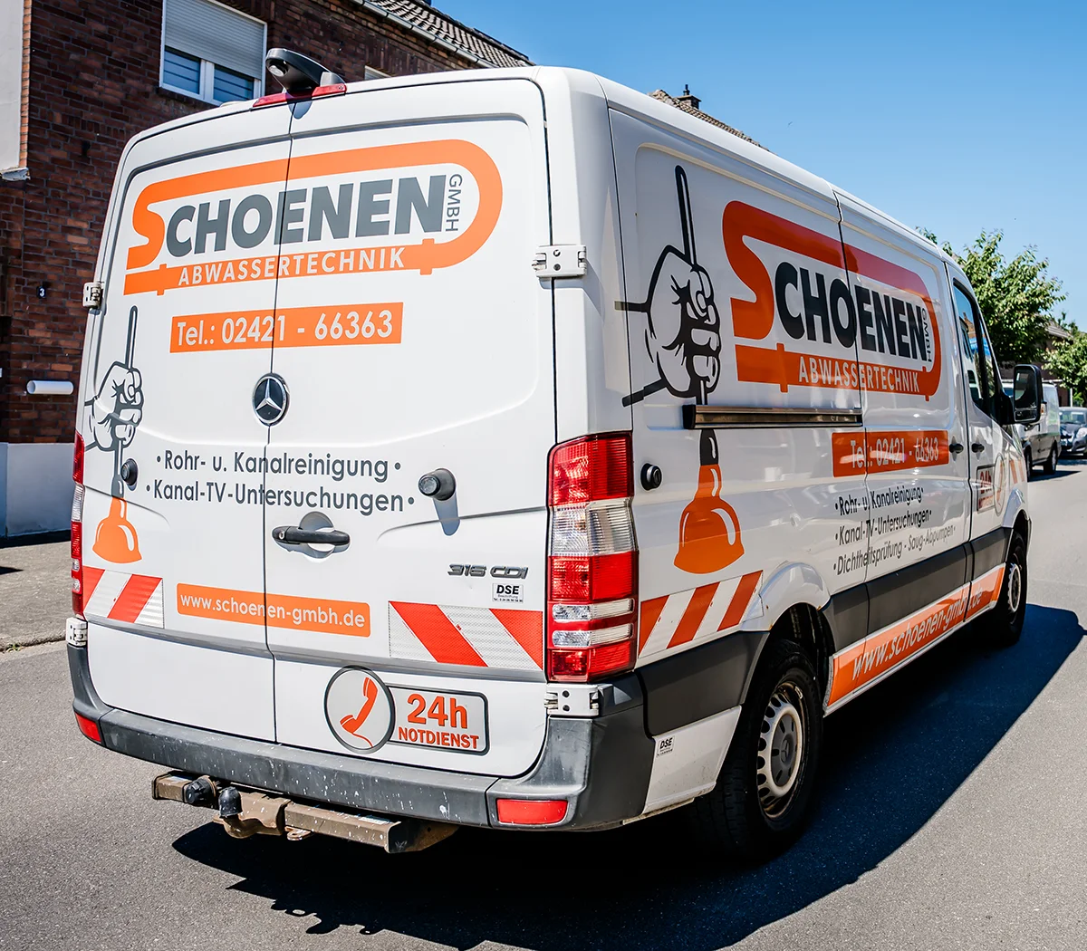 Ein weißer Servicewagen mit orangefarbener Aufschrift und dem Text "SCHOENEN Abwassertechnik" und einer Kolbenabbildung, geparkt an einer Straße in Düren. Der Wagen wirbt für Abflussreinigungs- und Rohrinspektionsdienste mit einer 24-Stunden-Notfall-Hotline.