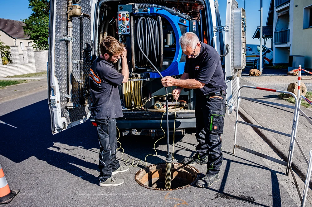 Zwei Mitarbeiter von Schoenen Abwassertechnik stehen an einer sonnigen Straße in Aachen an einem offenen Kanal neben einem Lieferwagen mit Ausrüstung und führen eine Kanalreinigung durch. Dabei führen Kabel und Werkzeuge in das Loch ein und scheinen sich auf Wartungsarbeiten an Abwasserkanälen oder Versorgungseinrichtungen zu konzentrieren.