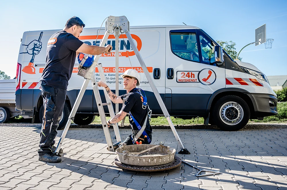Zwei Mitarbeiter stellen eine Stativausrüstung über einem offenen Kanal auf einer gepflasterten Fläche auf. Ein weißer Servicewagen mit orangefarbenen Akzenten und 24-Stunden-Notfalldienst-Schild ist an einem sonnigen Tag im Hintergrund geparkt.