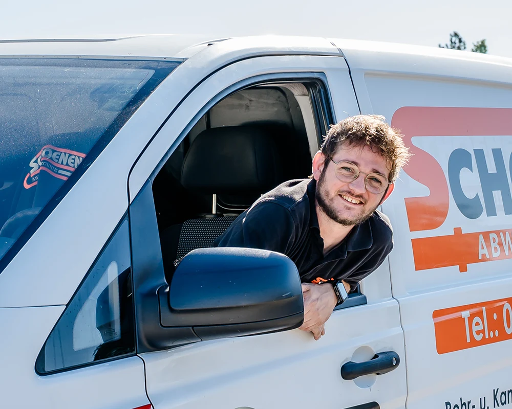 Ein lächelnder Mitarbeiter mit Brille von Schoenen Abwassertechnik lehnt sich an einem sonnigen Tag aus dem Fahrerfenster eines weißen Arbeitstransporters mit orange-roter Aufschrift an der Seite und blickt in die Kamera.