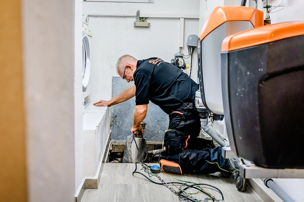 Ein Mitarbeiter von Schoenen Abwassertechnik in Arbeitskleidung kniet in einem Hauswirtschaftsraum auf dem Boden und arbeitet an freiliegenden Rohren und Geräten in Düren. Werkzeuge und eine große Maschine stehen in der Nähe, während der Klempner sich auf die Wartung konzentriert.