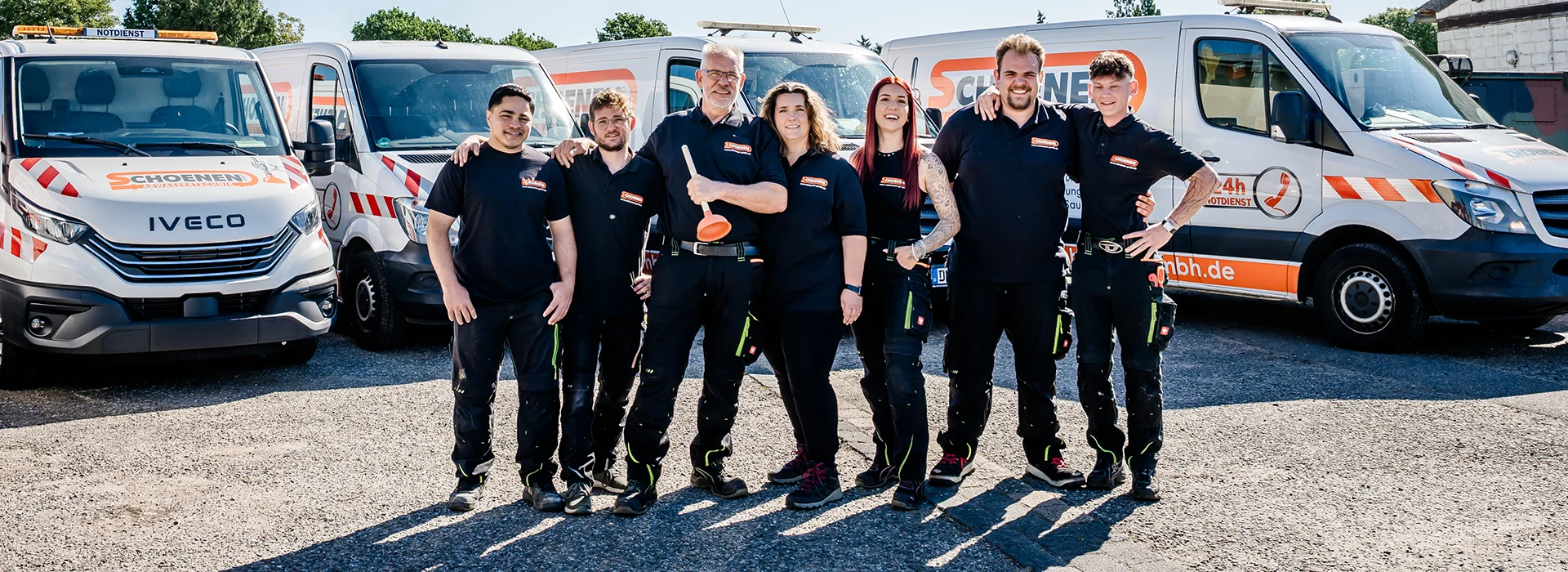 Das Team von Schoenen Abwassertechnik aus Düren in passenden dunklen Uniformen steht lächelnd vor drei Firmenfahrzeugen mit orangefarbenen und weißen Logos und posieren selbstbewusst an einem sonnigen Tag im Freien.